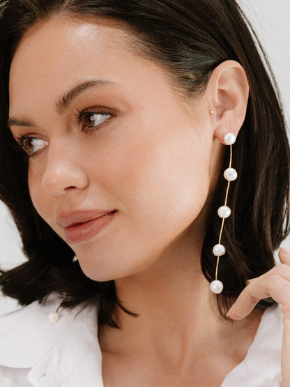 A woman with dark, shoulder-length hair wears a long, elegant earring featuring several pearls. She gently touches her ear and gazes thoughtfully to the side. Soft lighting enhances the delicate details.