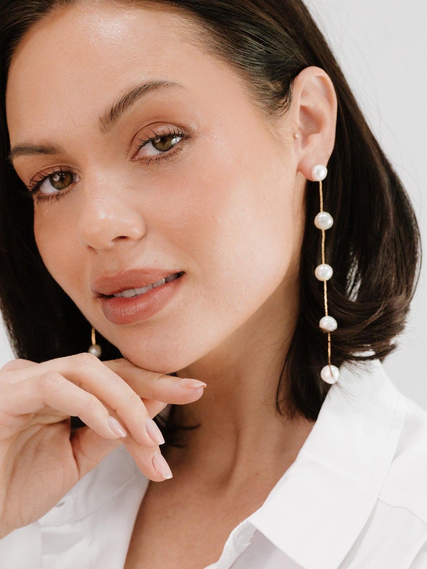 A woman with shoulder-length dark hair touches her chin, showcasing dangling pearl earrings. She has a subtle smile, wearing a white shirt against a soft, neutral background.