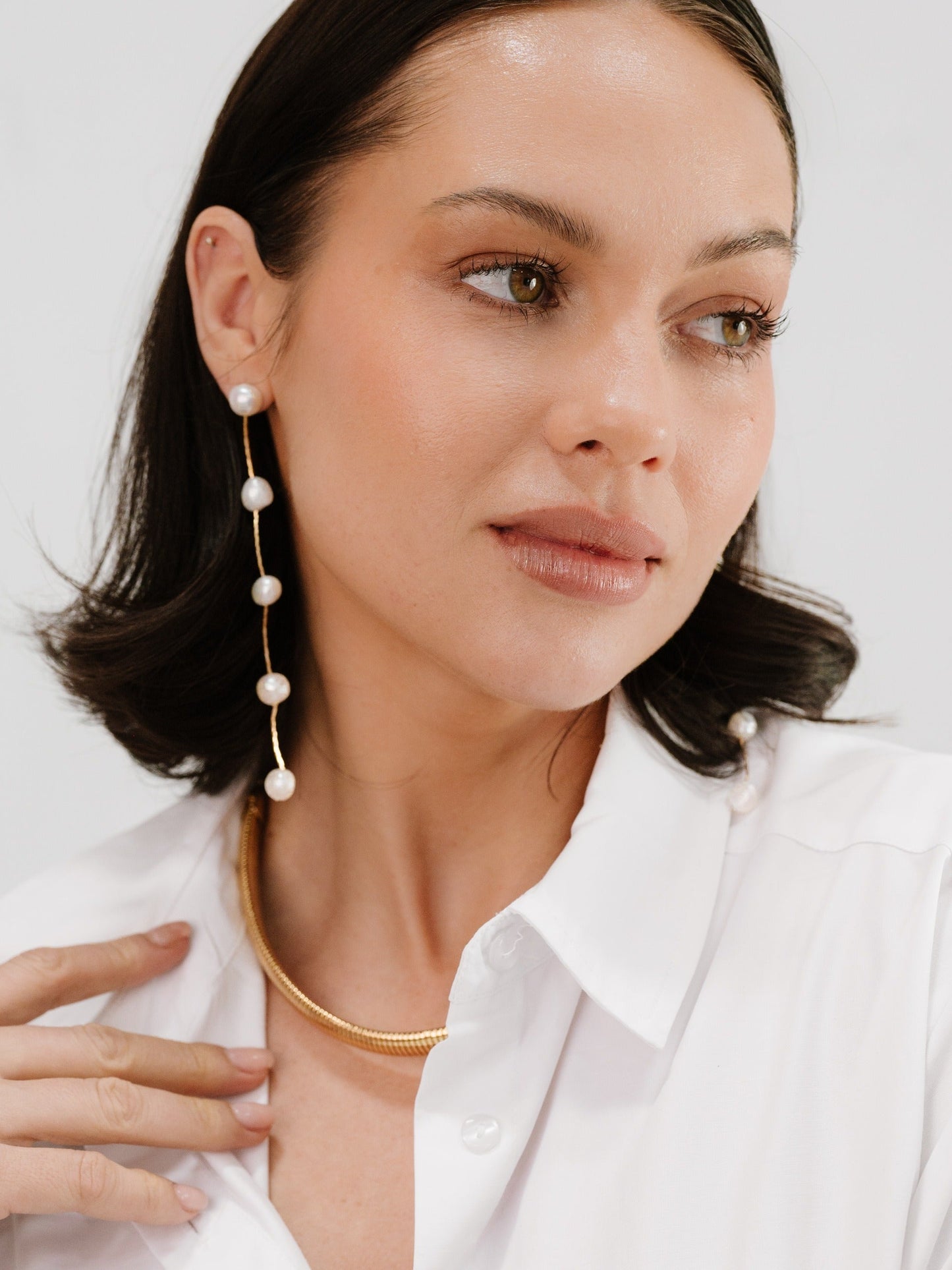 A woman with medium-length black hair, wearing a white shirt, poses gracefully. She has gold and pearl jewelry, highlighting her features against a neutral background, exuding elegance and style.