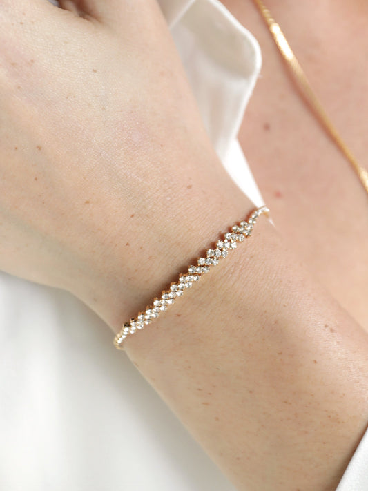 A delicate gold bracelet adorned with small sparkling stones is worn on a fair-skinned wrist. The hand rests against a light-colored fabric, suggesting a casual or elegant setting.