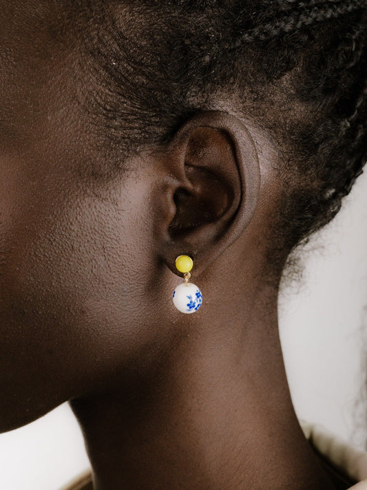 A close-up showcases an ear adorned with a colorful earring featuring a yellow circular top and a white spherical bottom decorated with blue floral patterns against dark skin and a light background.