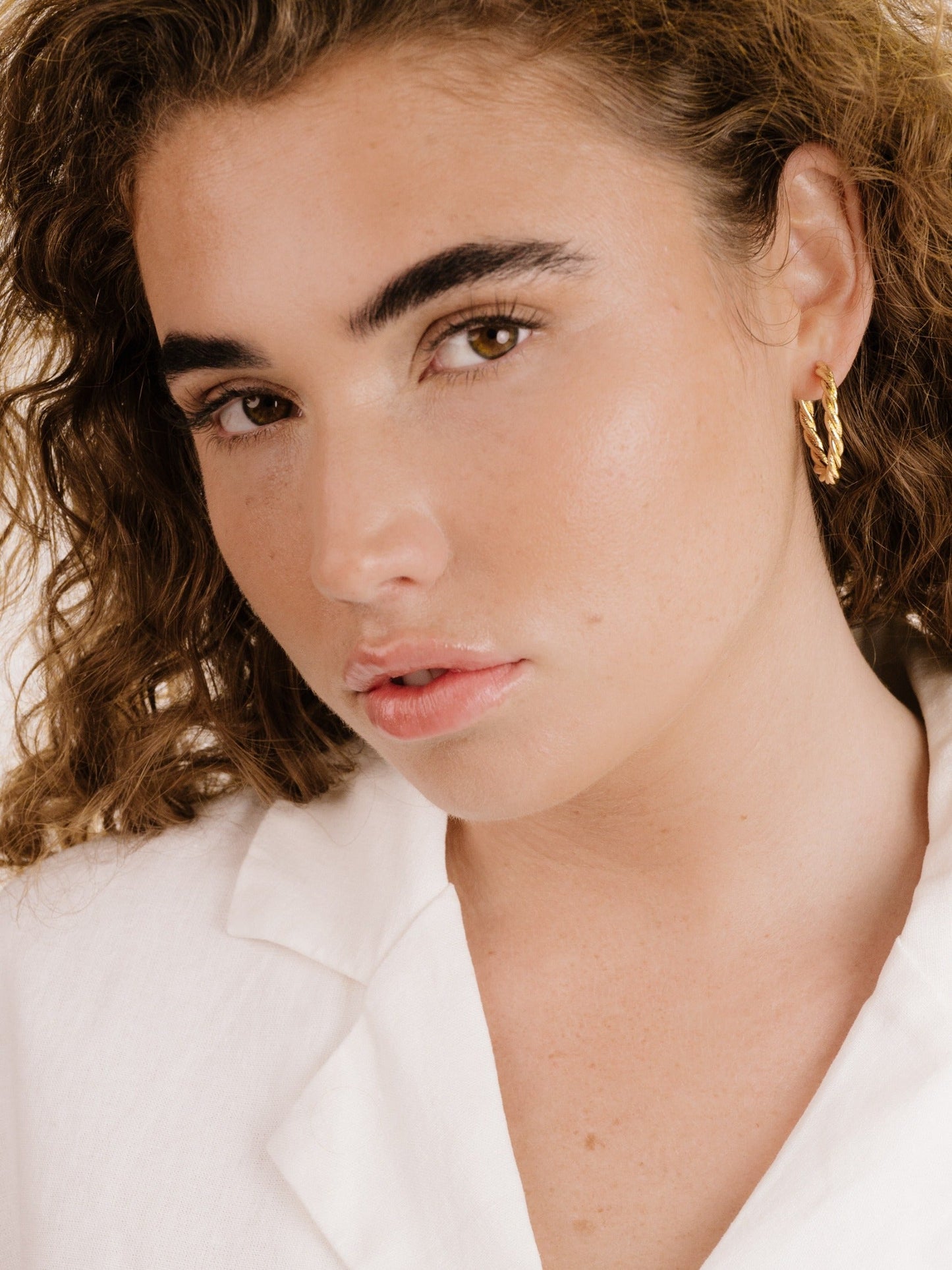 A young woman with curly hair gazes directly at the viewer, wearing a white shirt and a gold hoop earring, with a soft, subtle makeup look against a neutral background.