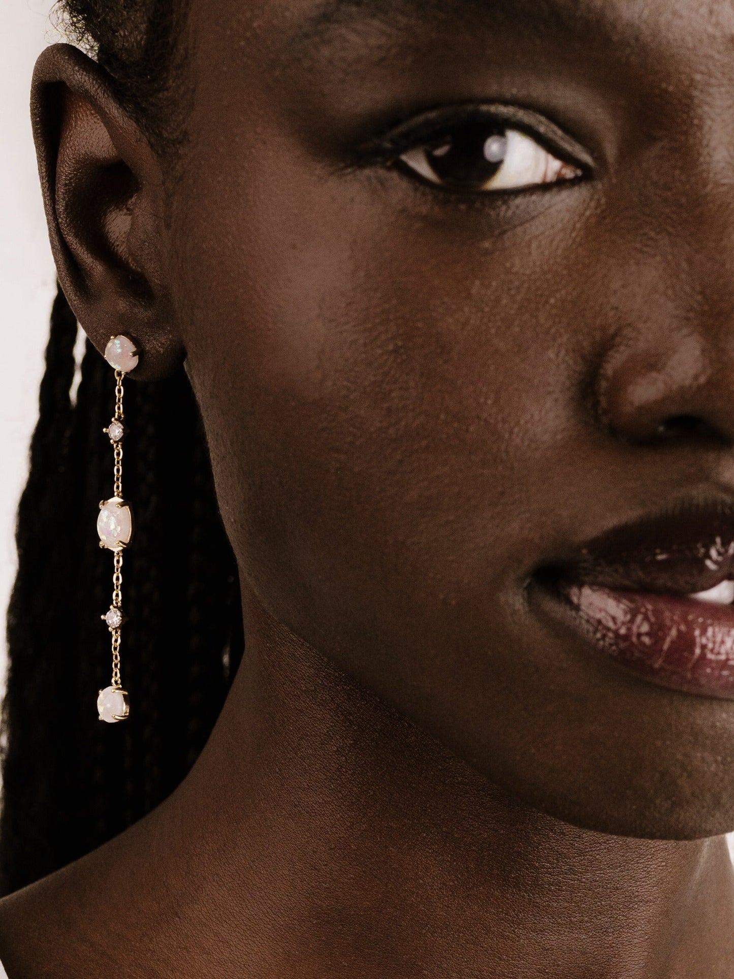 A close-up of a person's face showcases an elegant gold earring with three opalescent stones hanging from the earlobe. The skin tone is deep and rich, complementing the jewelry.