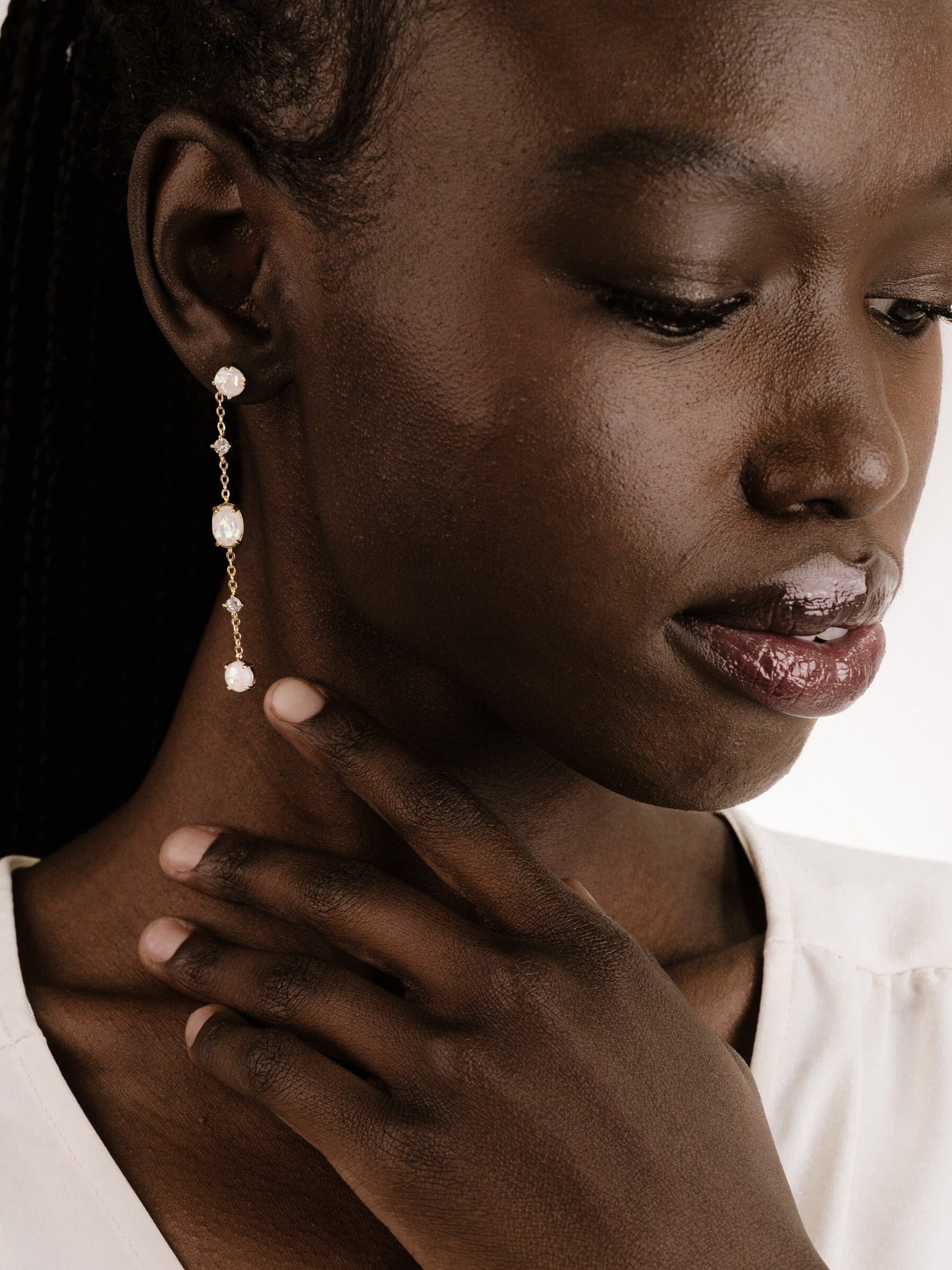 A person with dark skin wears a delicate, dangling earring featuring pearls and crystals. They gently touch their neck, showcasing the adornment against a light backdrop.