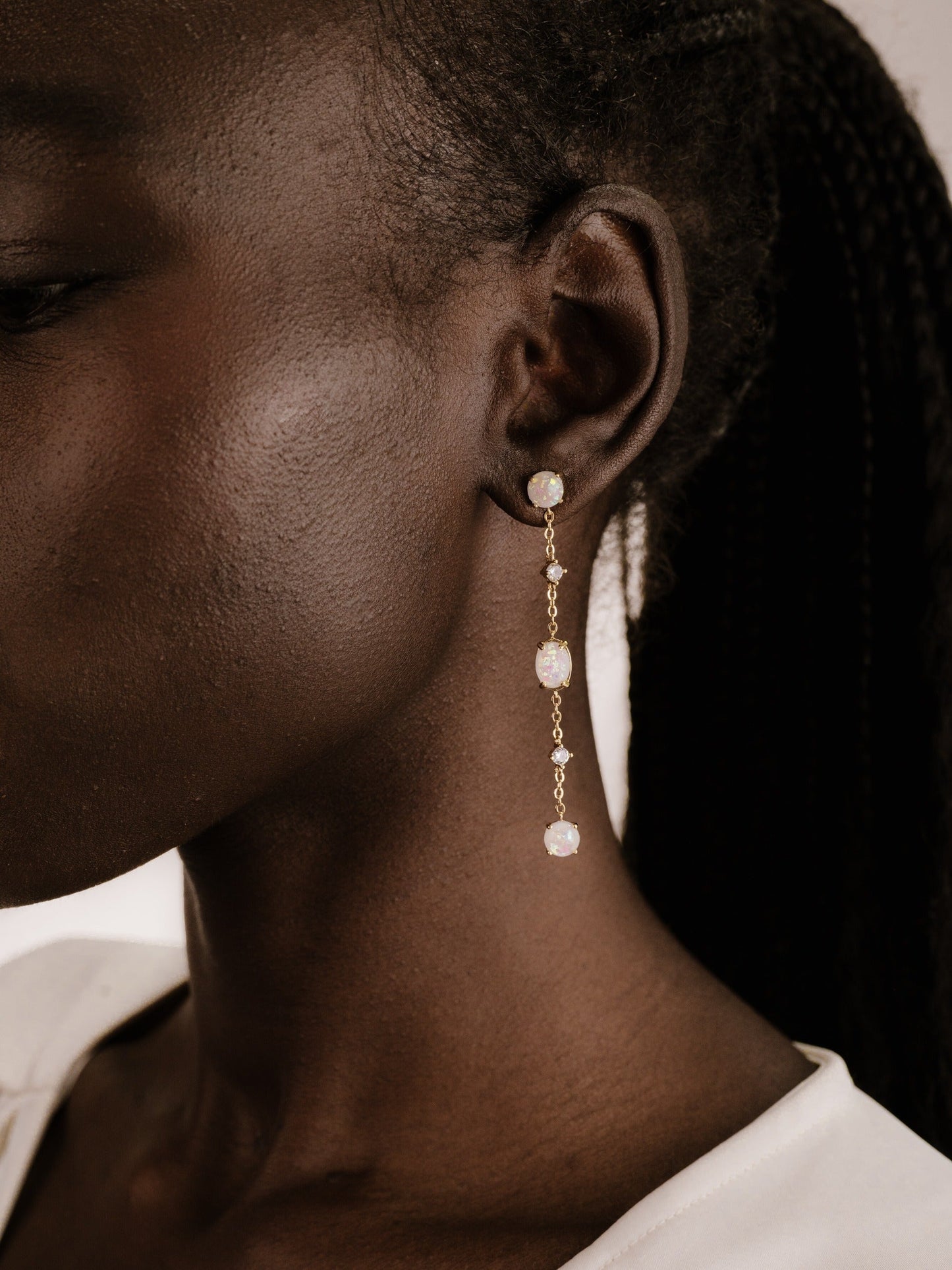 A close-up of a person's profile showcases an elegant, dangling earring with opalescent stones, accentuating the skin's texture against a softly blurred, neutral background.