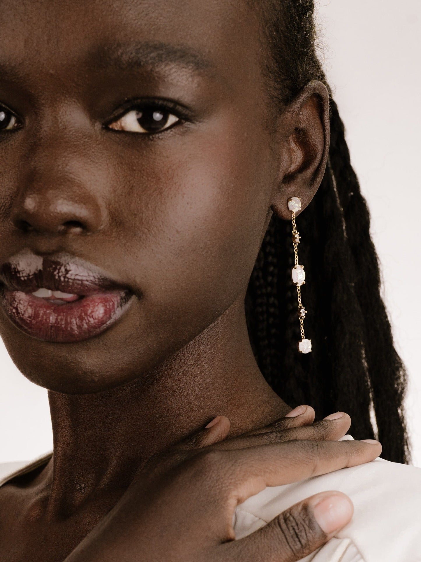 A close-up portrait of a person with dark skin, wearing elegant dangling earrings. They smile softly, with one hand resting on their shoulder, against a neutral background.