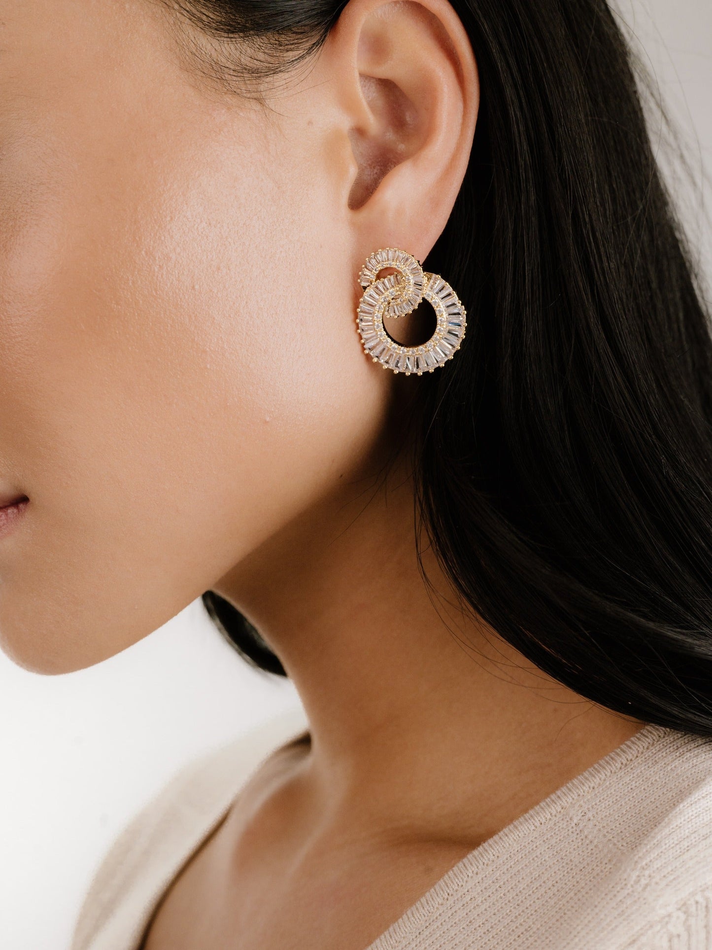 A close-up view of a woman's ear adorned with elegant, intertwined gold hoop earrings. The background is soft and neutral, highlighting the jewelry and the woman's smooth complexion.