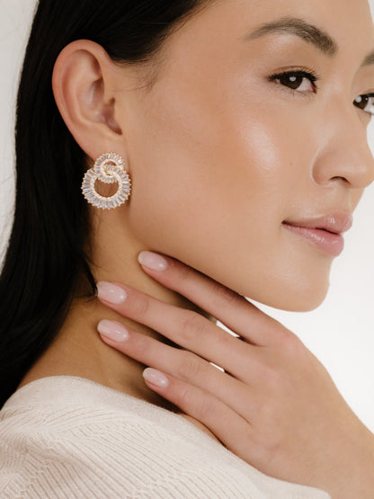 A woman with long black hair and smooth skin wears a decorative earring while gently touching her neck with her fingers, set against a neutral background.