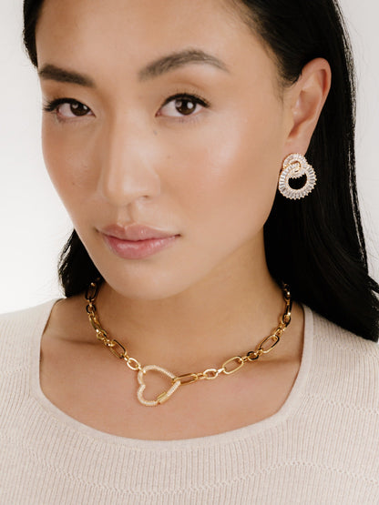 A woman models elegant jewelry, featuring large, textured earrings and a heart-shaped necklace, against a soft, neutral background that highlights the accessories and her warm complexion.