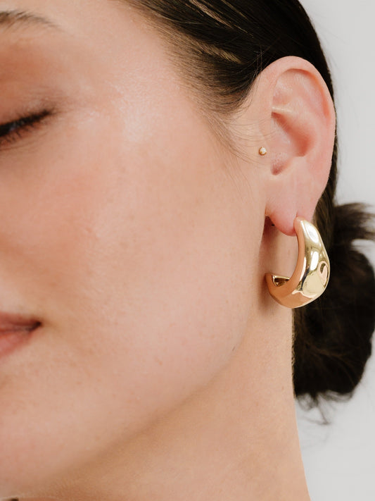 A close-up of a woman's face shows her with a relaxed expression. She wears a large, shiny gold hoop earring on one ear and has a small stud earring above it.