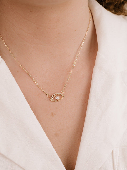 A gold necklace featuring an eye-shaped pendant adorned with a central gemstone and surrounding small stones rests gently on the skin of a person wearing a white collared shirt.