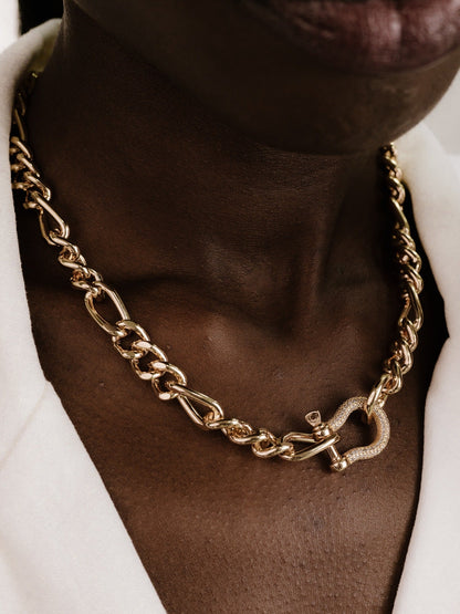 A gold chain necklace is displayed around a person's neck, featuring intertwined links and a decorative clasp, against a background of smooth, dark skin and a white garment.