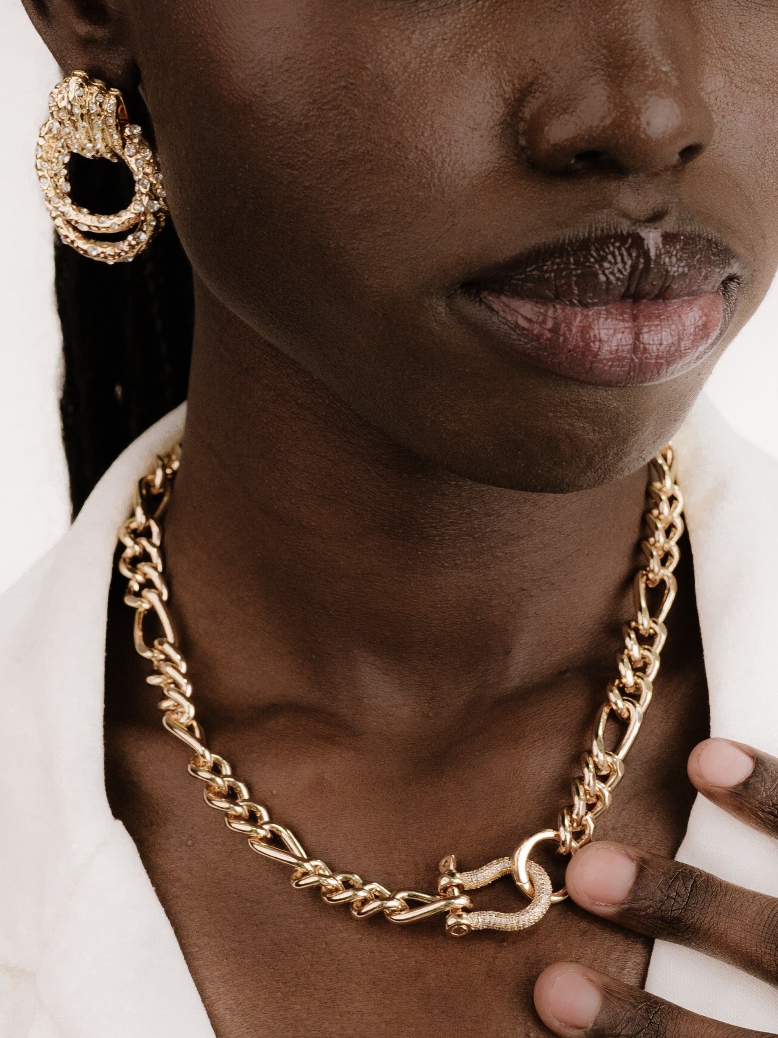 A close-up of a dark-skinned individual wearing a chunky gold chain necklace and large gold hoop earrings, with one hand gently touching the necklace, against a light-colored background.