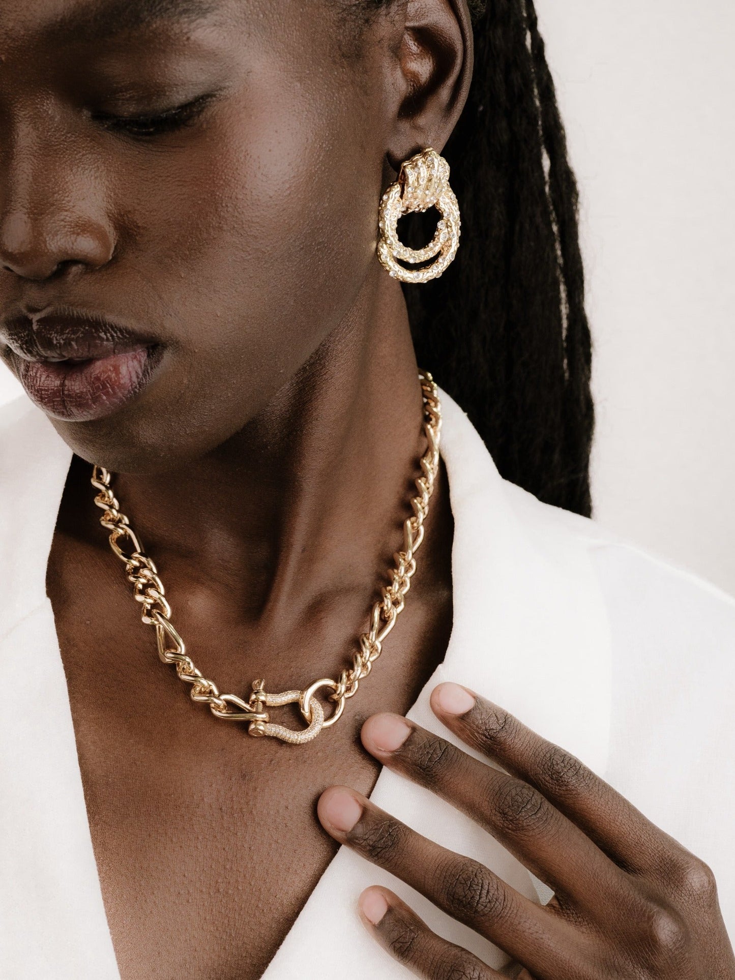 A person models gold jewelry, featuring large, intricate hoop earrings and a chunky chain necklace, while gently touching the necklace. They are against a neutral background, exuding elegance.