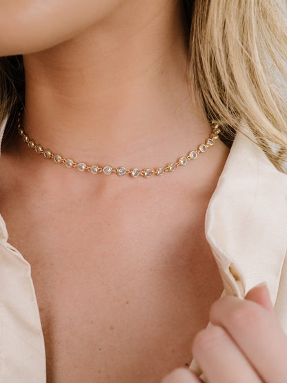 A gold choker necklace adorned with sparkling stones rests on the neck of a person, who is pulling at the collar of a light-colored shirt, suggesting a stylish or elegant context.