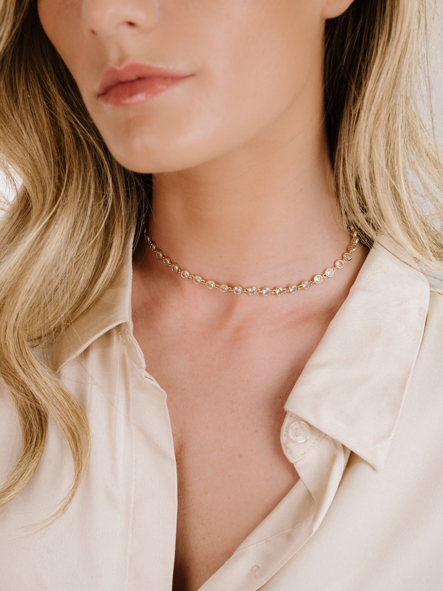 A close-up of a womanâ€™s neck adorned with a delicate gold choker featuring circular embellishments, set against a softly lit background. The woman wears a light-colored, silky shirt.