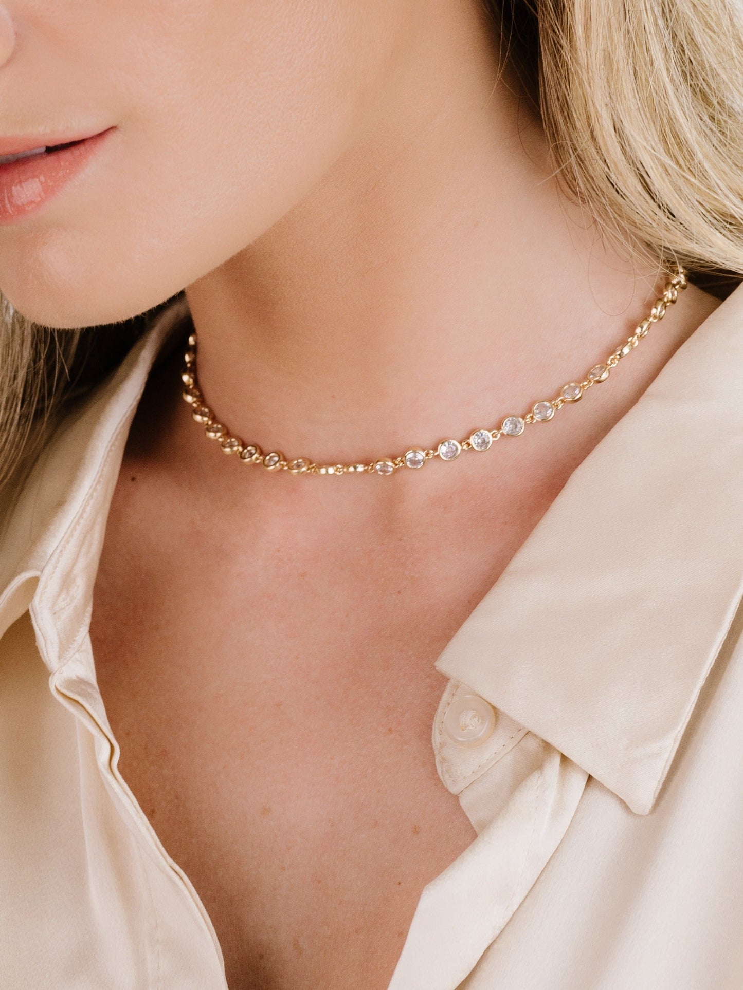 A close-up of a womanâ€™s neck showcases a delicate gold choker adorned with alternating beads and sparkling stones, against a backdrop of soft, cream-colored fabric.