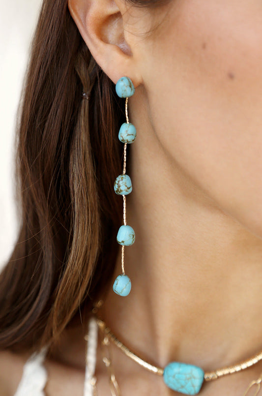 A turquoise bead earring hangs from the ear, featuring multiple beads arranged in a linear fashion. Nearby, a matching turquoise necklace adds to the jewelry ensemble.