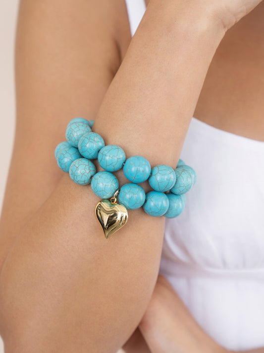 A turquoise beaded bracelet adorned with a golden heart charm embraces the wrist of a person in a white top, showcasing the accessory against a neutral background.