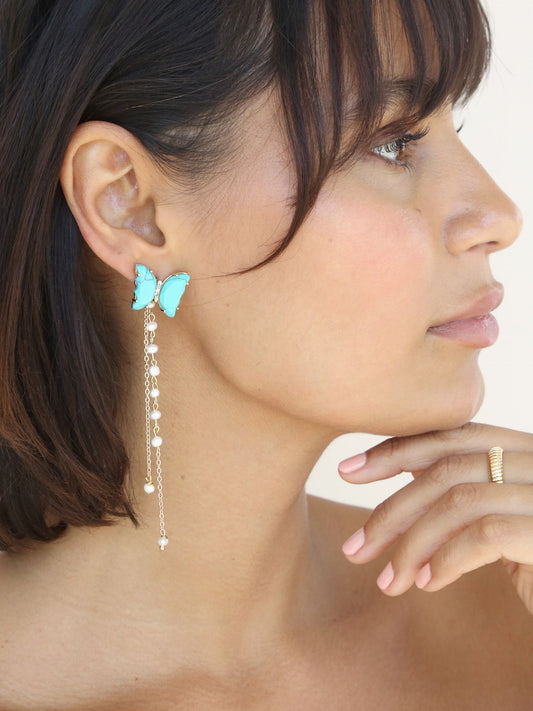 A woman is wearing a butterfly-shaped earring with dangling pearls, gently resting her chin on her hand. The background is neutral, enhancing her relaxed expression and stylish accessory.