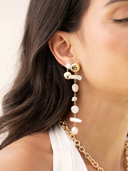 A gold and pearl earring dangles from a woman's ear, featuring a cascading design with various pearl shapes, set against a soft, neutral background and her long, dark hair.