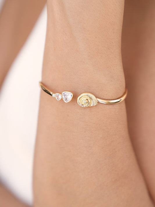 A thin gold bracelet features three decorative elements: a small diamond-like stone, a teardrop gem, and a spiral shell design, resting against a skin-toned wrist.