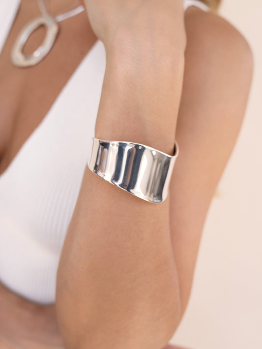 A shiny silver cuff bracelet is worn on a bare arm, highlighting its smooth, wavy surface under soft, neutral lighting in a minimalistic setting.