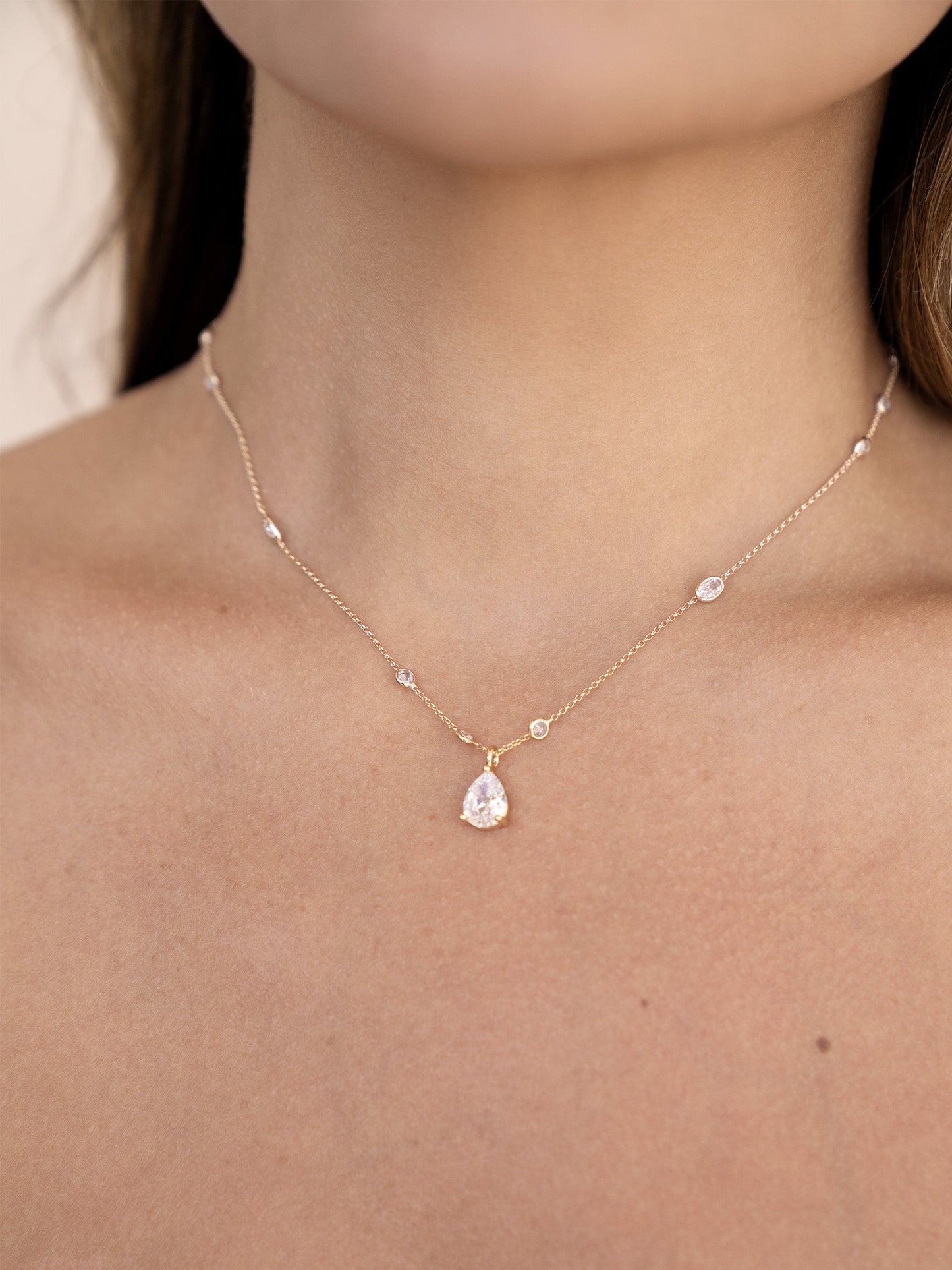 A delicate gold necklace adorns a bare neck, featuring a central pear-shaped diamond pendant with smaller diamond accents, set against a softly blurred, light background.