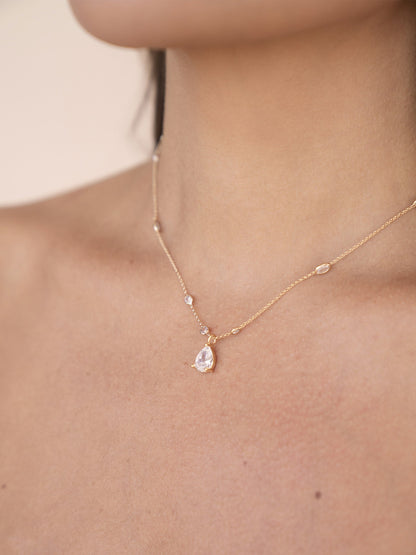 A delicate gold necklace rests on a bare neck, featuring a pear-shaped pendant and small gemstones, set against a soft, neutral background that enhances its elegance.