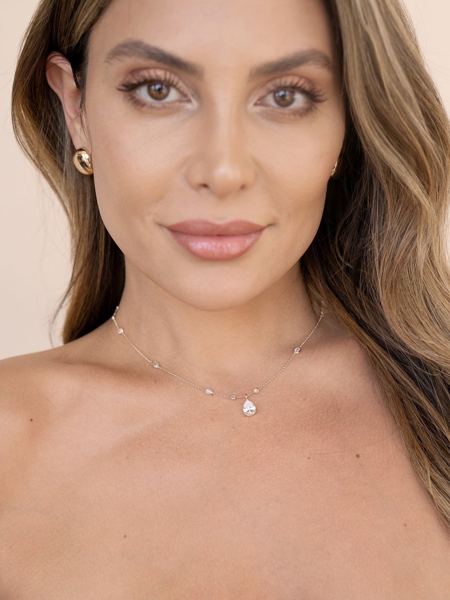 A woman with long, wavy hair is looking directly at the camera, wearing a delicate silver necklace with a teardrop pendant. She has natural makeup and gold earrings, against a soft beige background.