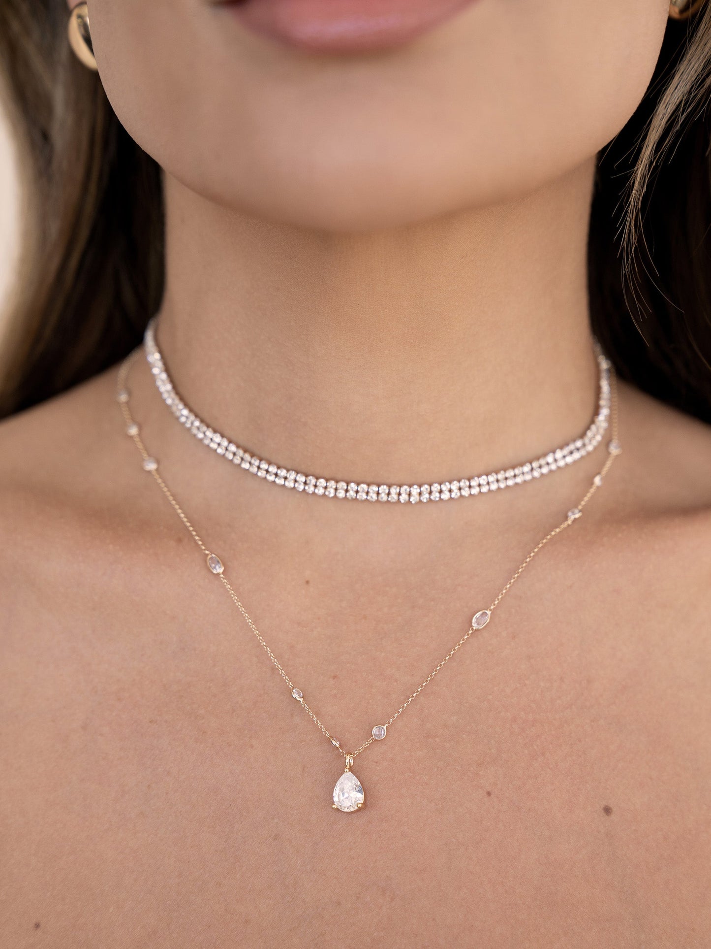 A close-up of a person's neck showcases two delicate necklaces. One is a sparkling choker, while the other features a teardrop pendant, enhancing the elegance against the bare skin.