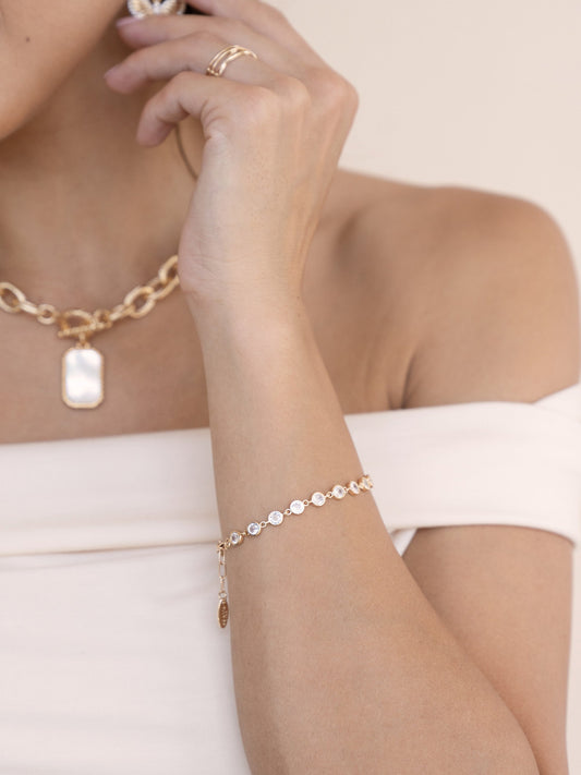 A hand adorned with a delicate gold bracelet featuring circular stones adjusts a pendant necklace. The subject is wearing a strapless top and is set against a neutral background.