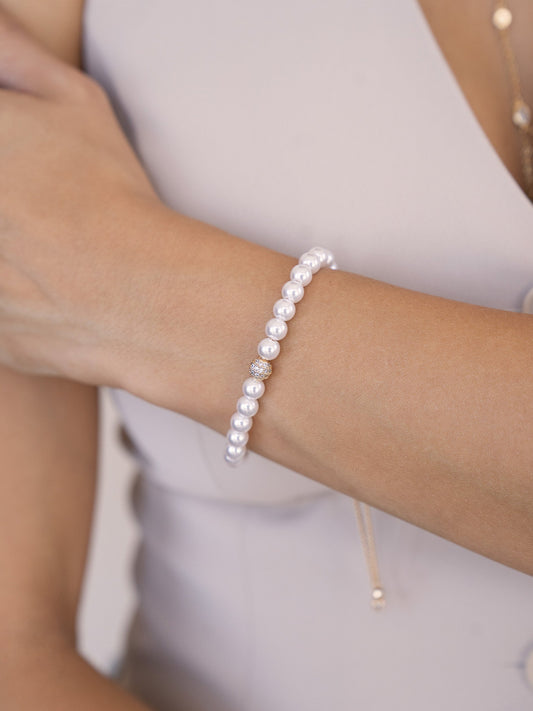 A pearl bracelet adorned with small circular pearls and a shiny centerpiece is worn on a hand with a light skin tone, against a neutral background with a soft, elegant attire.