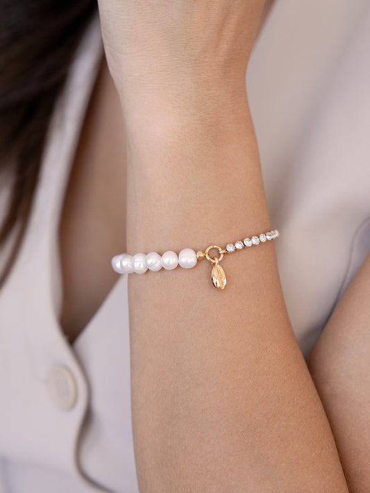 A bracelet made of alternating pearls and sparkling gems adorns a person's wrist. The wrist is exposed, with a blurred background of light fabric and skin, suggesting a casual setting.