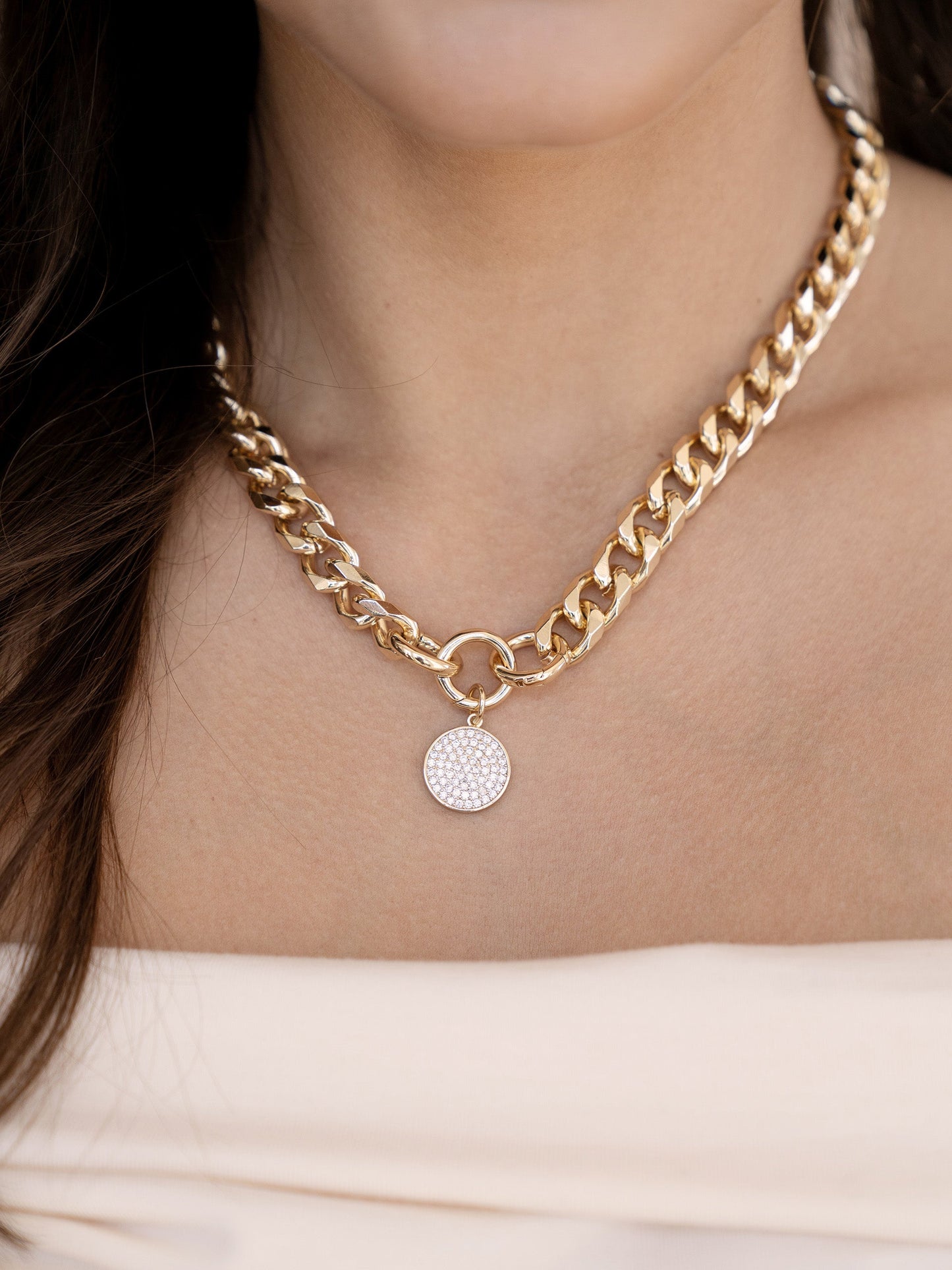 A gold chain necklace adorns a person's neck, featuring interlinked large links and a circular pendant with a textured surface, set against soft, neutral fabric.