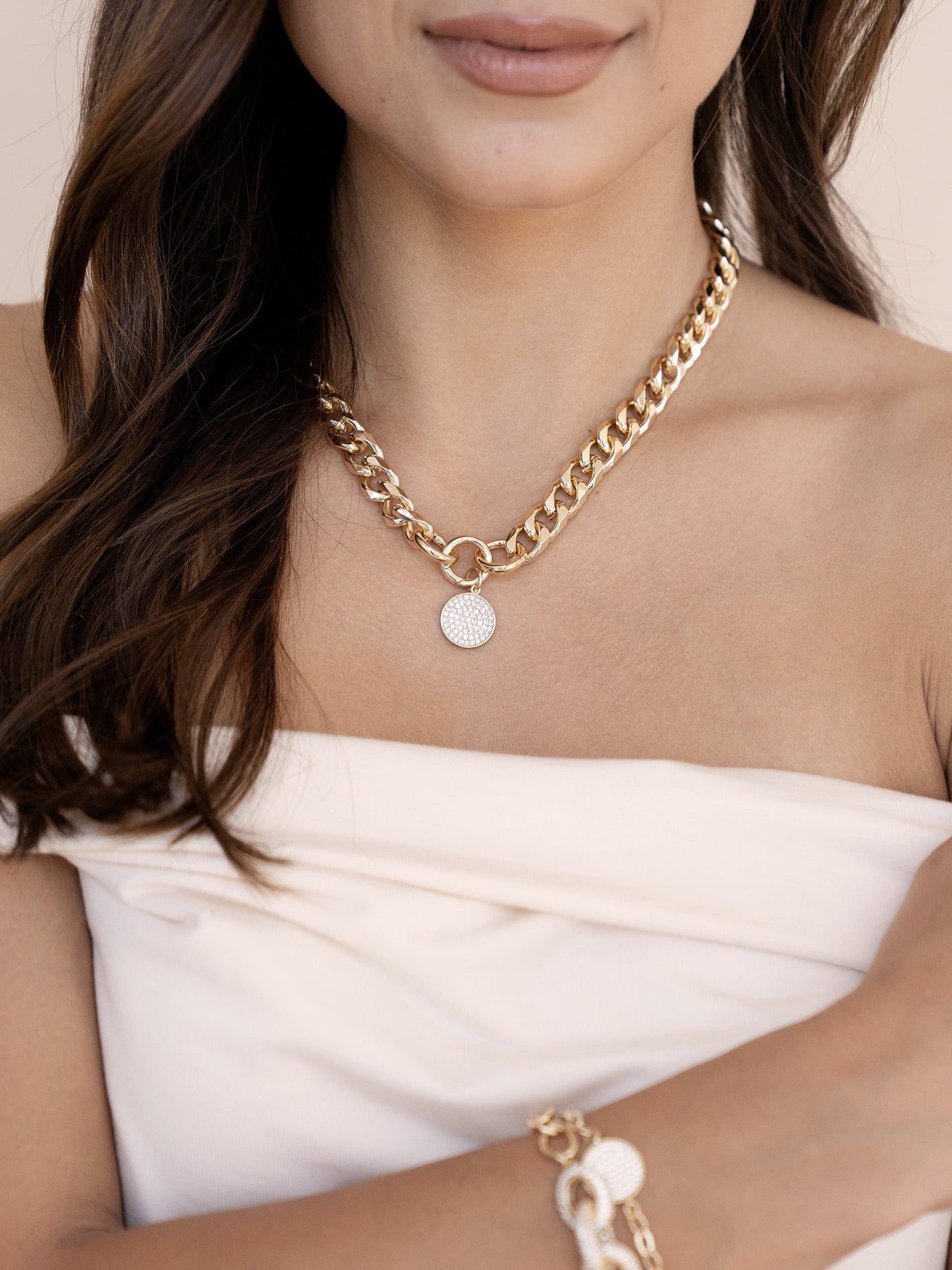 A woman wearing a gold chain necklace with a circular pendant smiles softly. She is dressed in a strapless top, with her hair cascading over her shoulder, set against a neutral background.