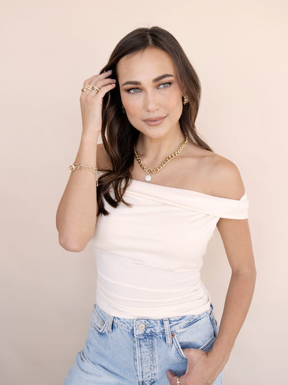 A woman with long, dark hair stands, gently touching her hair with one hand. She wears a light off-shoulder top and high-waisted jeans, set against a soft beige background.