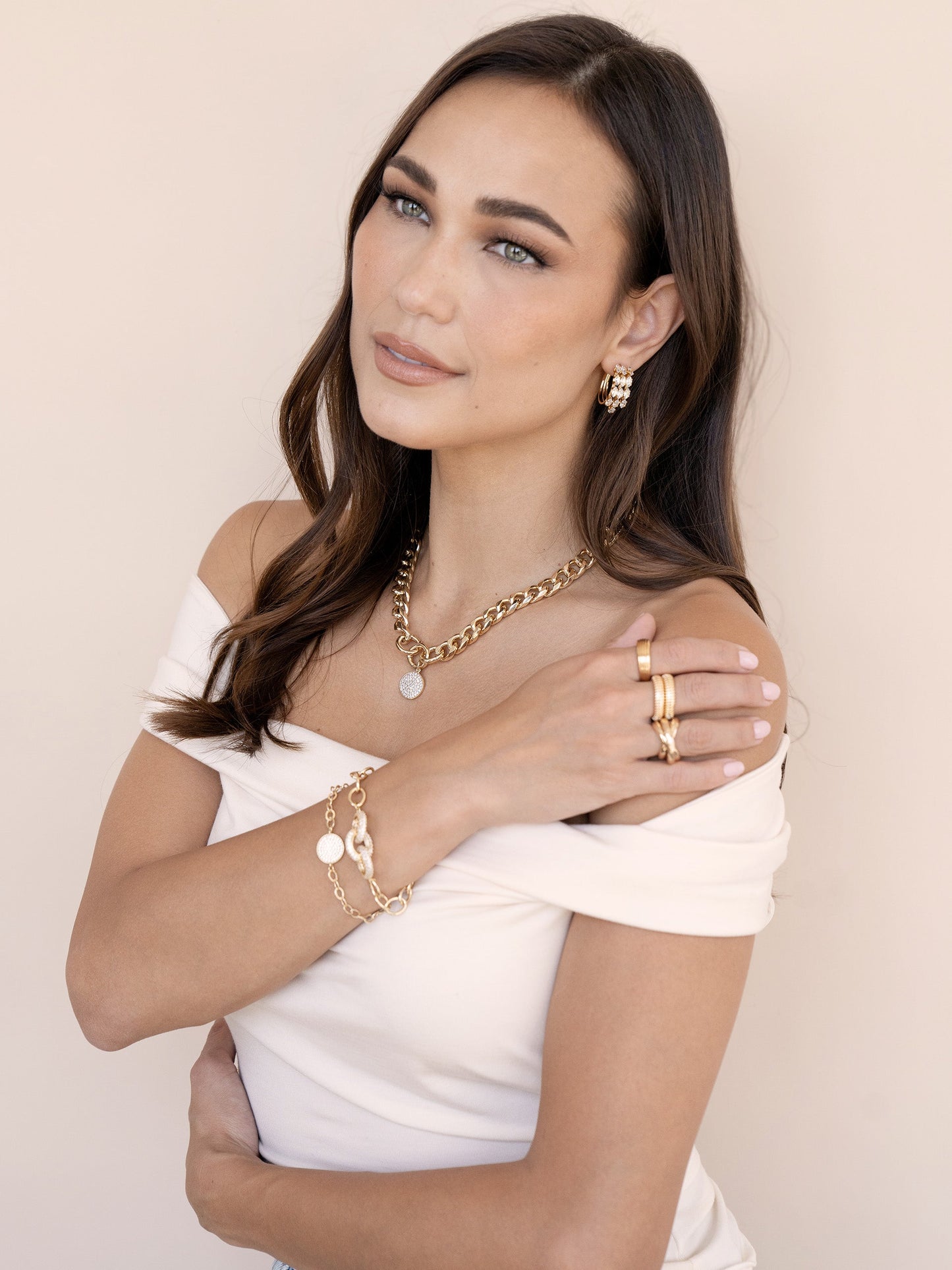 A woman with long, flowing hair poses gracefully, dressed in a white off-shoulder top adorned with various gold jewelry, set against a soft beige background.