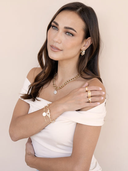 A woman with long, flowing hair poses gracefully, dressed in a white off-shoulder top adorned with various gold jewelry, set against a soft beige background.