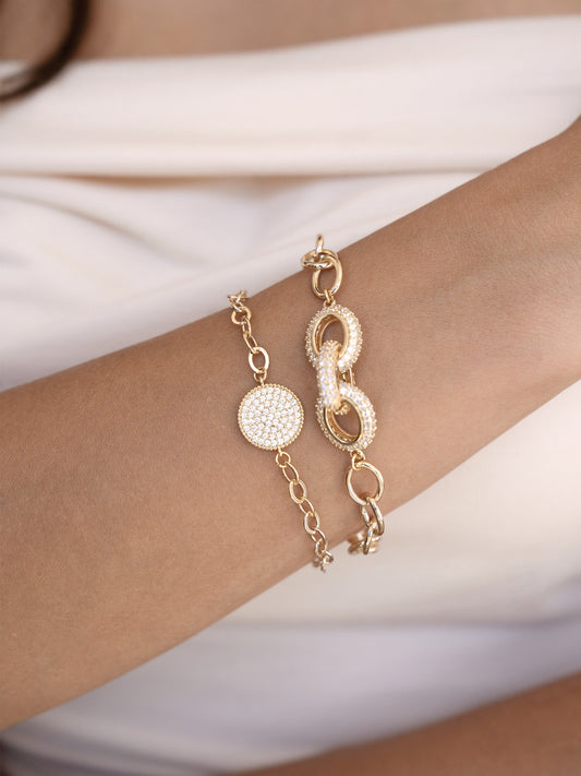 Two gold bracelets adorn a wrist; one is decorated with a circular, sparkling disk, while the other features interlinked, shiny oval shapes. They rest against a light fabric background.