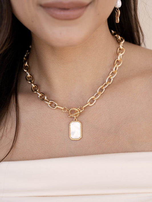 A gold chain necklace with a rectangular pendant hangs on a woman's neck, complemented by her soft lips. She wears a strapless top against a minimal background.