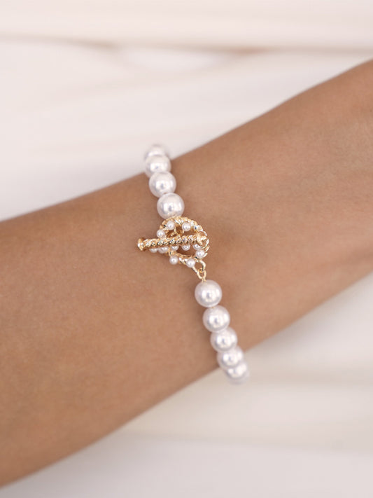 A gold and pearl bracelet adorns a wrist, featuring alternating white pearls and a decorative, intricate gold clasp. The skin is set against a soft, light-colored background.