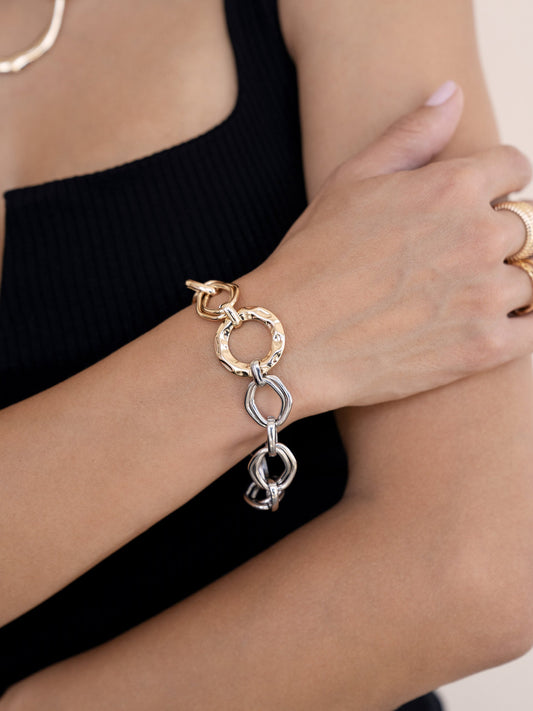 A hand wearing a stylish metal bracelet is shown. The bracelet features interconnected gold and silver links. The background is neutral, enhancing the focus on the accessory.