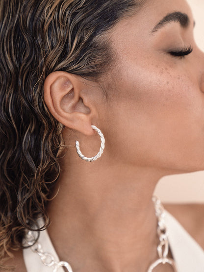 A close-up of a person's ear adorned with a twisted silver hoop earring, as they pose with their eyes closed, showcasing natural, wavy hair and subtle makeup against a light background.