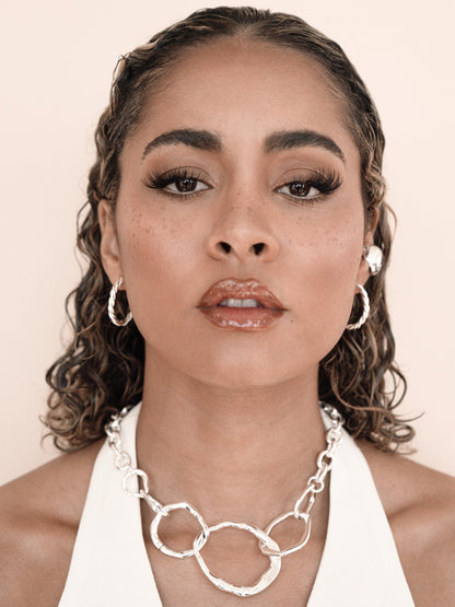 A woman with curly hair gazes directly at the viewer, wearing bold makeup and silver jewelry, including earrings and a chunky necklace, against a soft, neutral background.