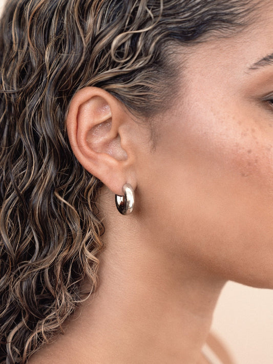 A close-up of a woman's ear adorned with a shiny silver hoop earring, with loose, wet, wavy hair cascading around her shoulder, emphasizing a natural and relaxed appearance.