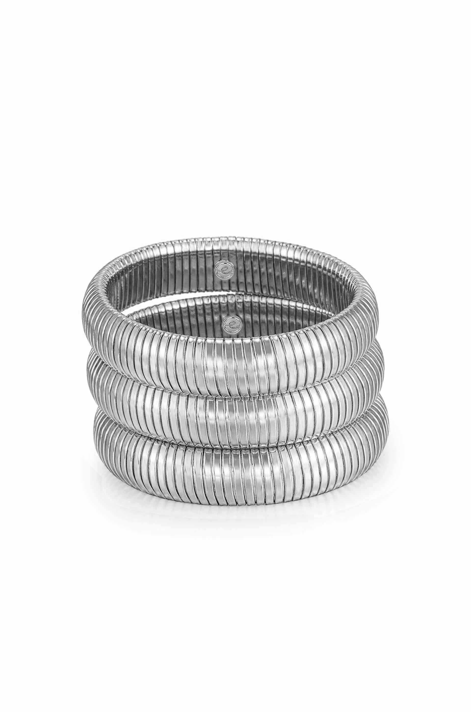 Three interconnected, polished metal bangles rest on a smooth surface, showcasing their coiled design and shiny finish, reflecting light in a minimalist setting.