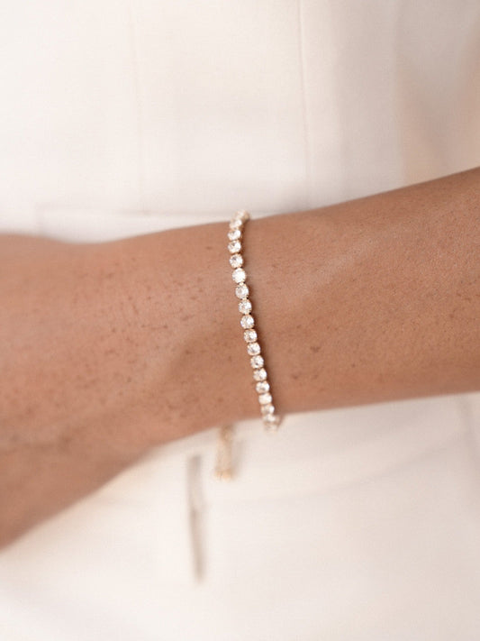 A gold bracelet adorned with sparkling gemstones encircles a wrist. The skin tone is warm against the minimalist backdrop, emphasizing the elegance of the jewelry and the personâ€™s attire.