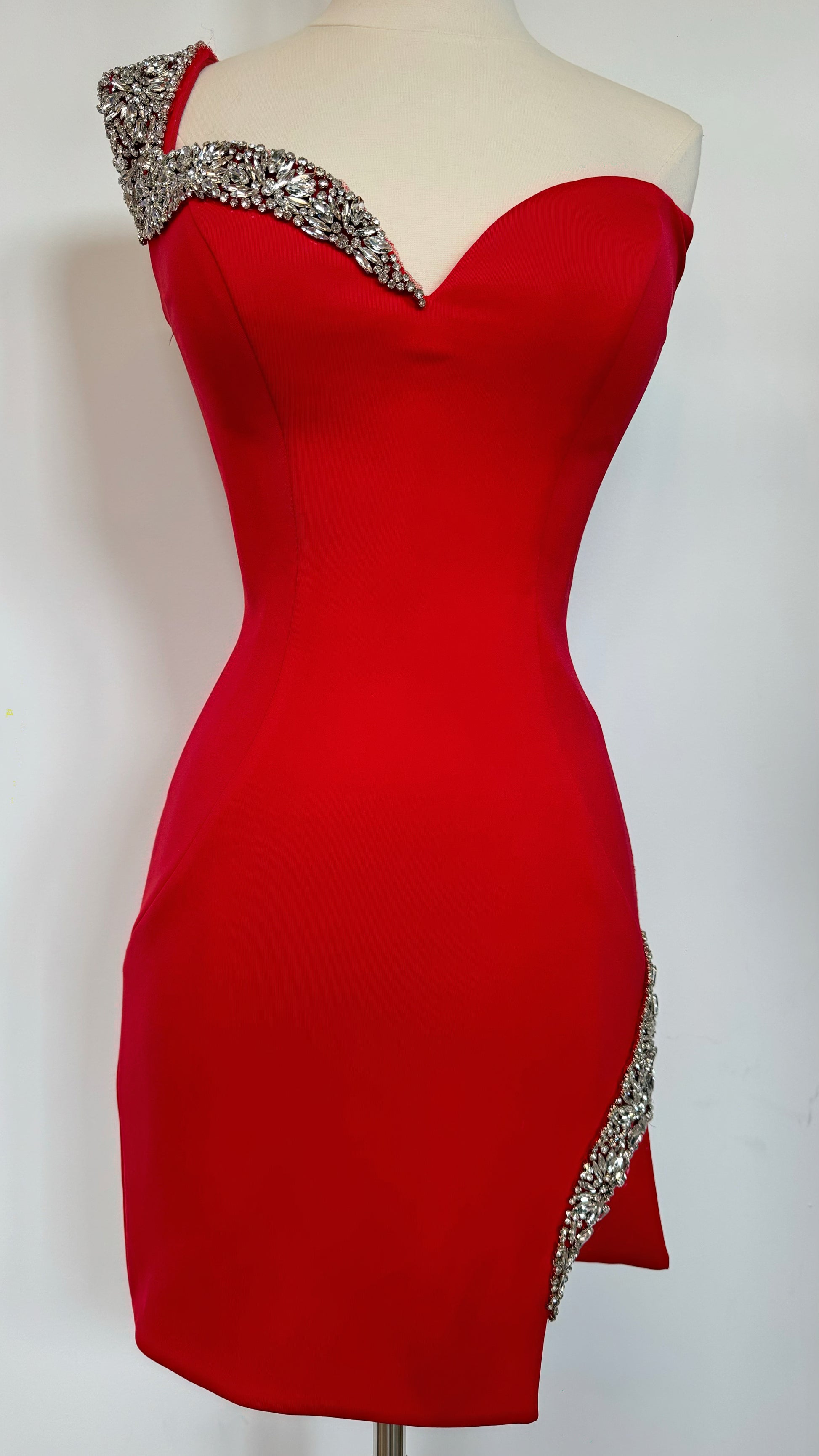 A vibrant red dress is displayed on a mannequin. The dress features an asymmetric neckline adorned with a sparkling embellishment on one shoulder and a decorative trim along the side.