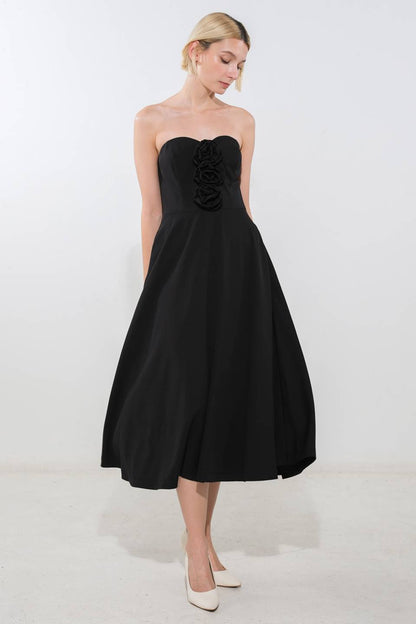 A woman stands gracefully in a strapless black dress featuring three fabric roses at the chest. She is in a minimalistic bright space with a light-colored floor.